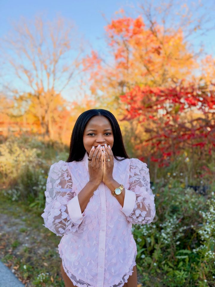 Frida in a pink lace shirt, laughing behind her hands, with autumn trees behind her